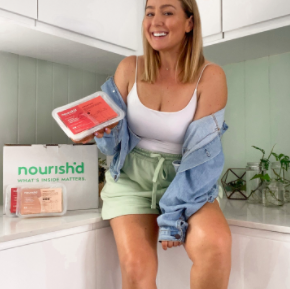 Instagram image of a young woman sitting at a small dining table  eating her Nourish'd fresh meal from a bow, with a crystal glass of kombucha beside her.