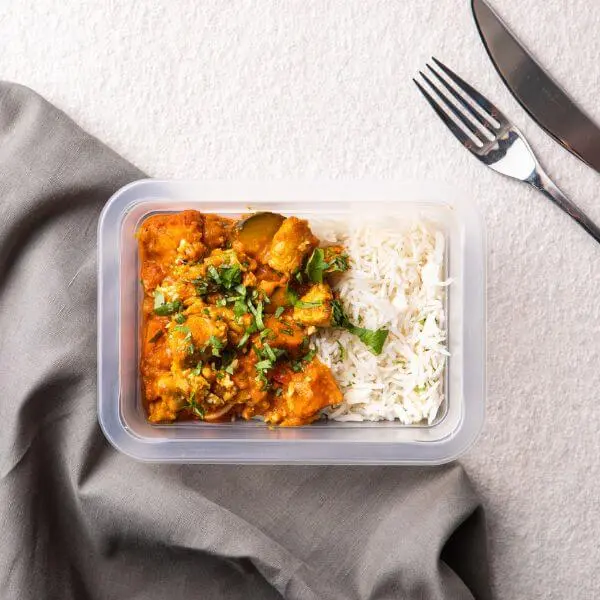Clear container shot of the Butter Chicken with Basmati Rice, perfectly portioned and ready to go. The container is placed on the grey countertop with the dark grey cloth nearby. The parsley garnish and creamy texture of the dish shine through, emphasizing its fresh, homemade appeal.
