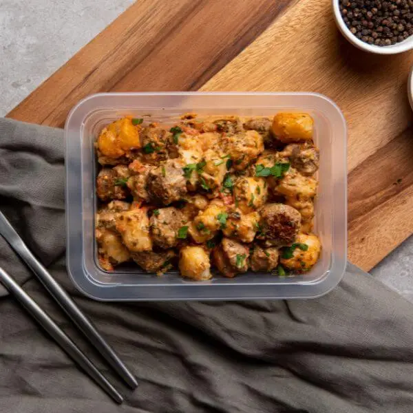A top down photograph of an Italian Sausage Gnocchi served in a container, set upon a wooden chopping board and dark grey cloth. A knife and fork sits to the left, and some peppercorns sit in a ramekin to the right.