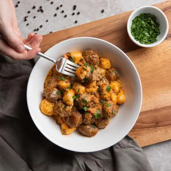 A top down photograph of a hand holding a fork, digging into a white bowl of Italian Sausage Gnocchi as if about to eat. The bowl is set upon a wooden chopping board and dark grey cloth, with peppercorns and a ramekin of chopped green herbs on the side.