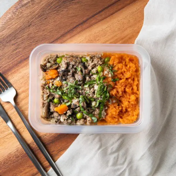 A clear plastic container of De-constructed Shepherd’s Pie garnished with chopped spring onions. The container is placed on a wooden cutting board with a beige cloth visible underneath. A fork and knife are positioned to the left, ready to dig into the hearty meal.