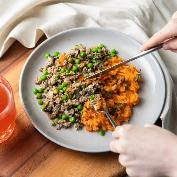 An action shot of De-constructed Shepherd’s Pie. One hand holds a knife, and the other holds a fork, cutting into the dish. A red juice cup in a glass adds a pop of color in the background. The dish, garnished with spring onions, is presented on a wooden cutting board with a beige cloth underneath, all on a grey marble surface.