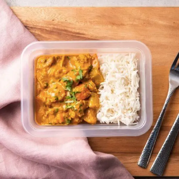 A clear plastic container of Chicken Masala with Basmati Rice, showcasing the creamy Masala-spiced curry paired with perfectly cooked basmati rice. Garnished with vibrant chopped parsley, the container is placed on a wooden chopping board. A fork and knife rest to the right, with a pink cloth adding a touch of warmth to the scene.