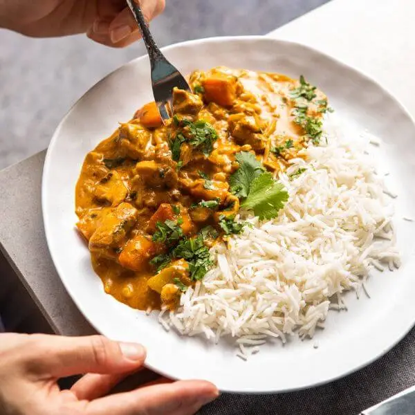 An action shot of Chicken Masala with Basmati Rice, showing a fork lifting a piece of tender, spiced chicken. The other hand holds the white plate, which features the creamy curry and fragrant basmati rice, all garnished with fresh parsley.