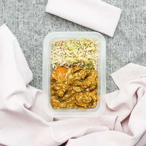 A clear plastic container of Chicken Masala with Cauliflower Rice, displaying the rich Indian-style gravy coating tender chicken and cauliflower rice. Chopped parsley is sprinkled on top as a garnish. The container is surrounded by a light pink cloth and a black and white patterned cloth, adding warmth and texture to the shot.