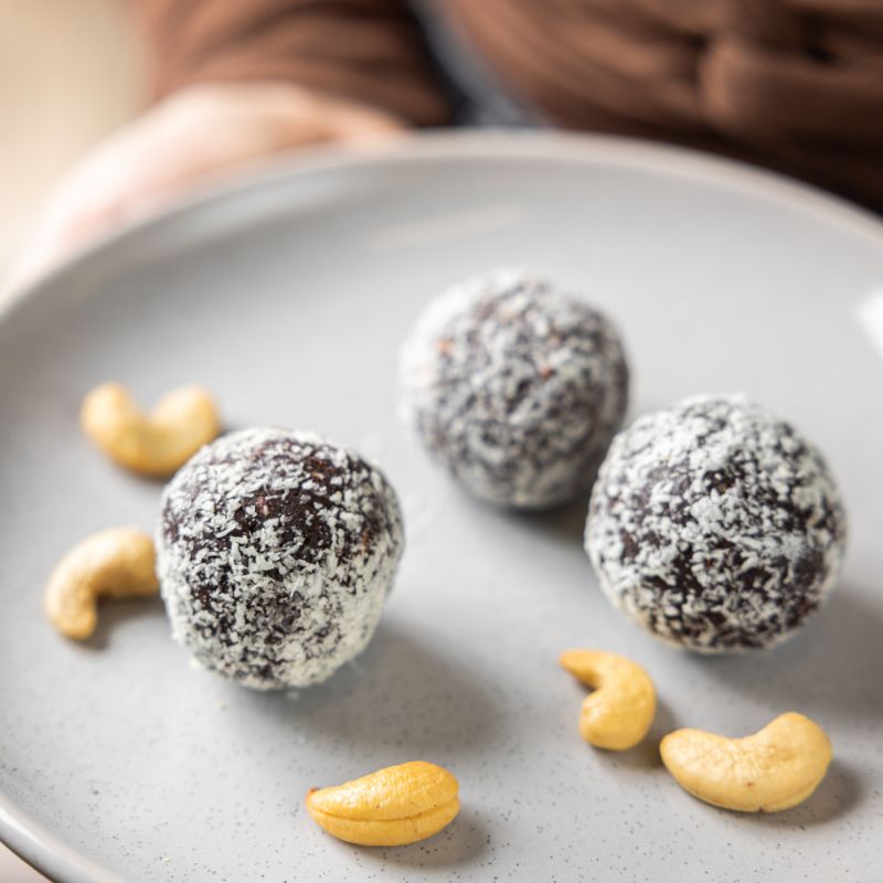 A close up of the three chocolate protein balls, dusted with dessicated coconut, on a white plate, with cashews to decorate the plate around it.