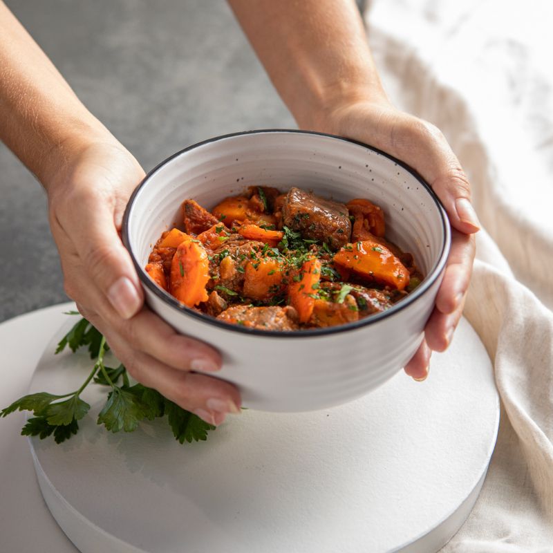 An action shot of two hands lifting a white bowl with a black rim, filled with Slow Cooked Beef Stew. The bowl rests on a white saucer plate, and a cream-colored cloth sits underneath. The dark grey marble counter and parsley leaves provide a fresh and rustic background