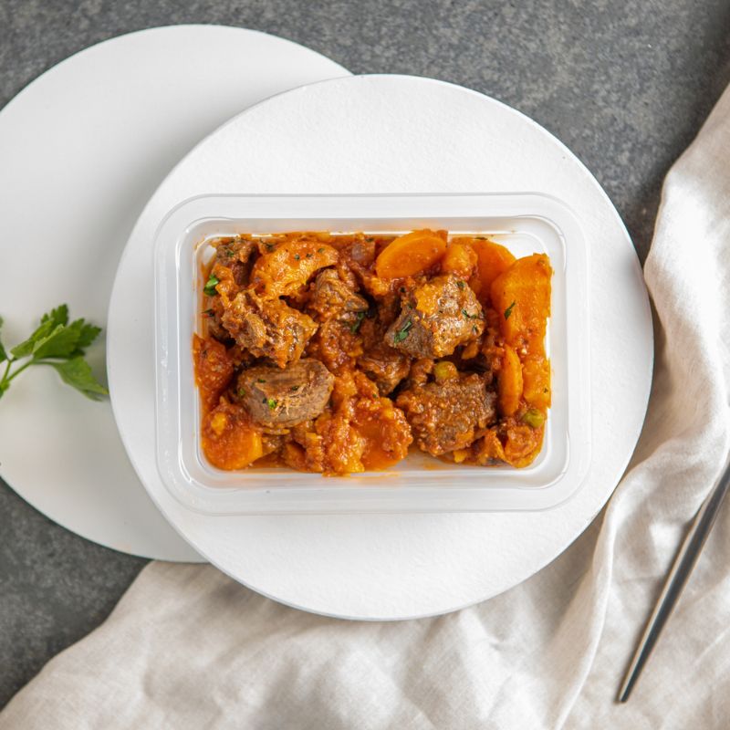 A clear plastic container of Slow Cooked Beef Stew, sitting on a cream-colored cloth atop a dark grey marble counter. Garnished with chopped spring onion, the stew is packed with tender beef and root vegetables. Fresh parsley leaves are scattered as decorative elements.
