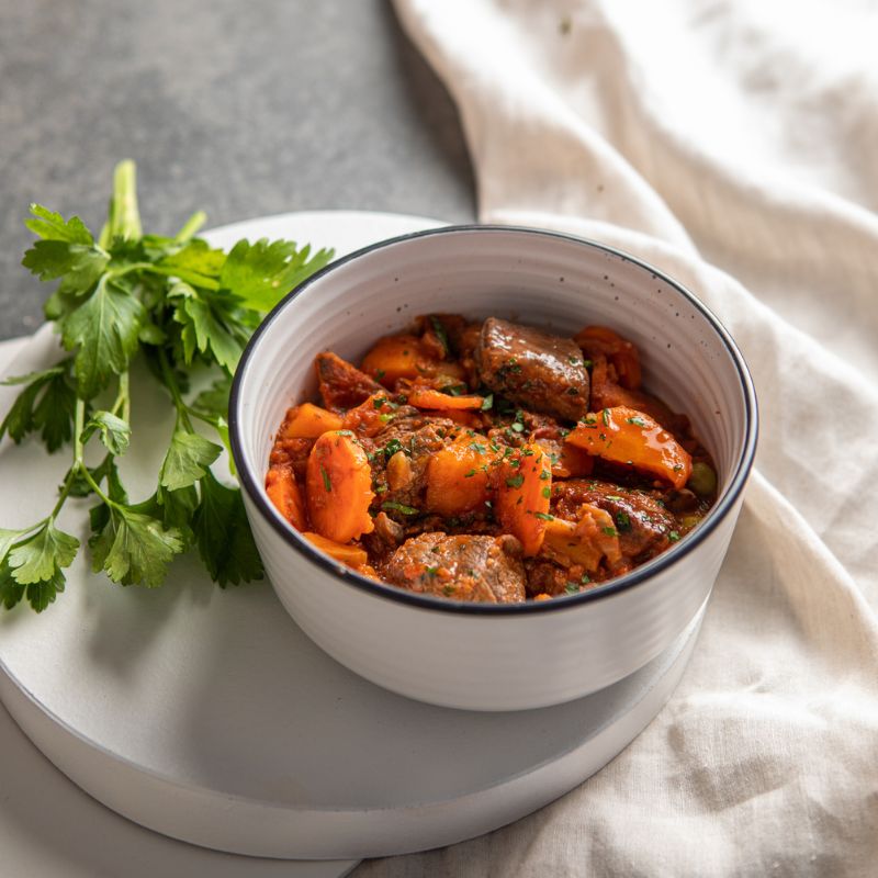 A side-angle view of Slow Cooked Beef Stew in a white bowl with a black rim. The bowl is placed on a white saucer plate with a cream-colored cloth tucked underneath. The stew is garnished with fresh chopped spring onion, with parsley leaves in the background.