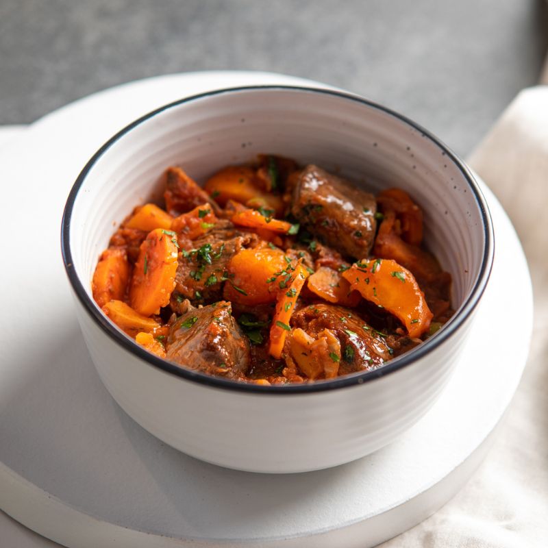 A close-up shot of Slow Cooked Beef Stew, highlighting its rich tomato base, tender chunks of grass-fed beef, and hearty root vegetables. Garnished with fresh chopped spring onion for a vibrant pop of color.