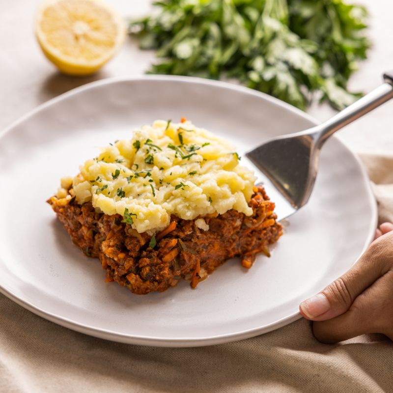 Action shot of a spatula digging into the creamy mashed potato topping of the Cottage Pie, showcasing its hearty filling underneath. The same beige-colored cloth and grey marble countertop elements are present for consistency.