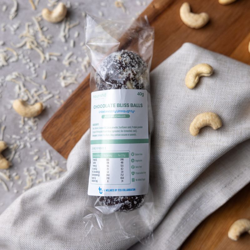 A top-down photograph of three chocolate protein balls wrapped in their packaging with a nutrition label and description on the front, placed on a tan cloth and a wooden chopping board, surround by cashews and a sprinkle of dessicated coconut.