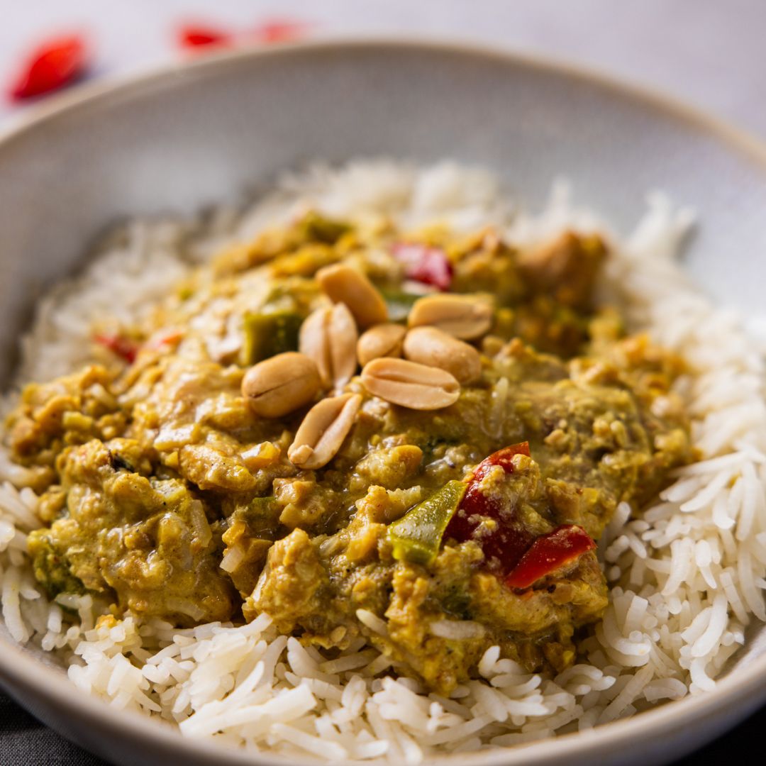 A close up photograph of a creamy, rich satay chicken with capsicum and peanuts on a bed of basmati rice in a ceramic bowl.