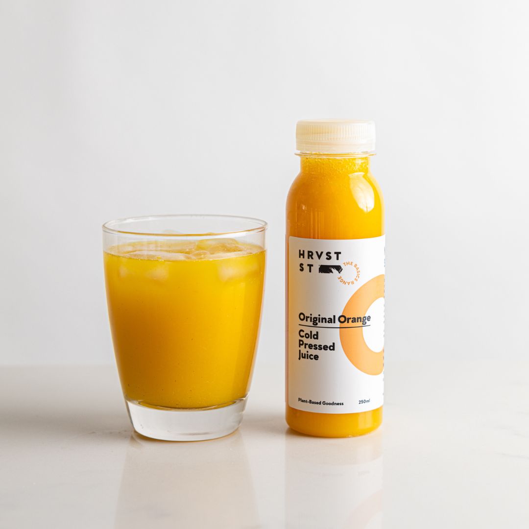 A drinking glass of orange juice, served beside a bottled serve of cold pressed orange juice, set against a white grey backdrop and bench.