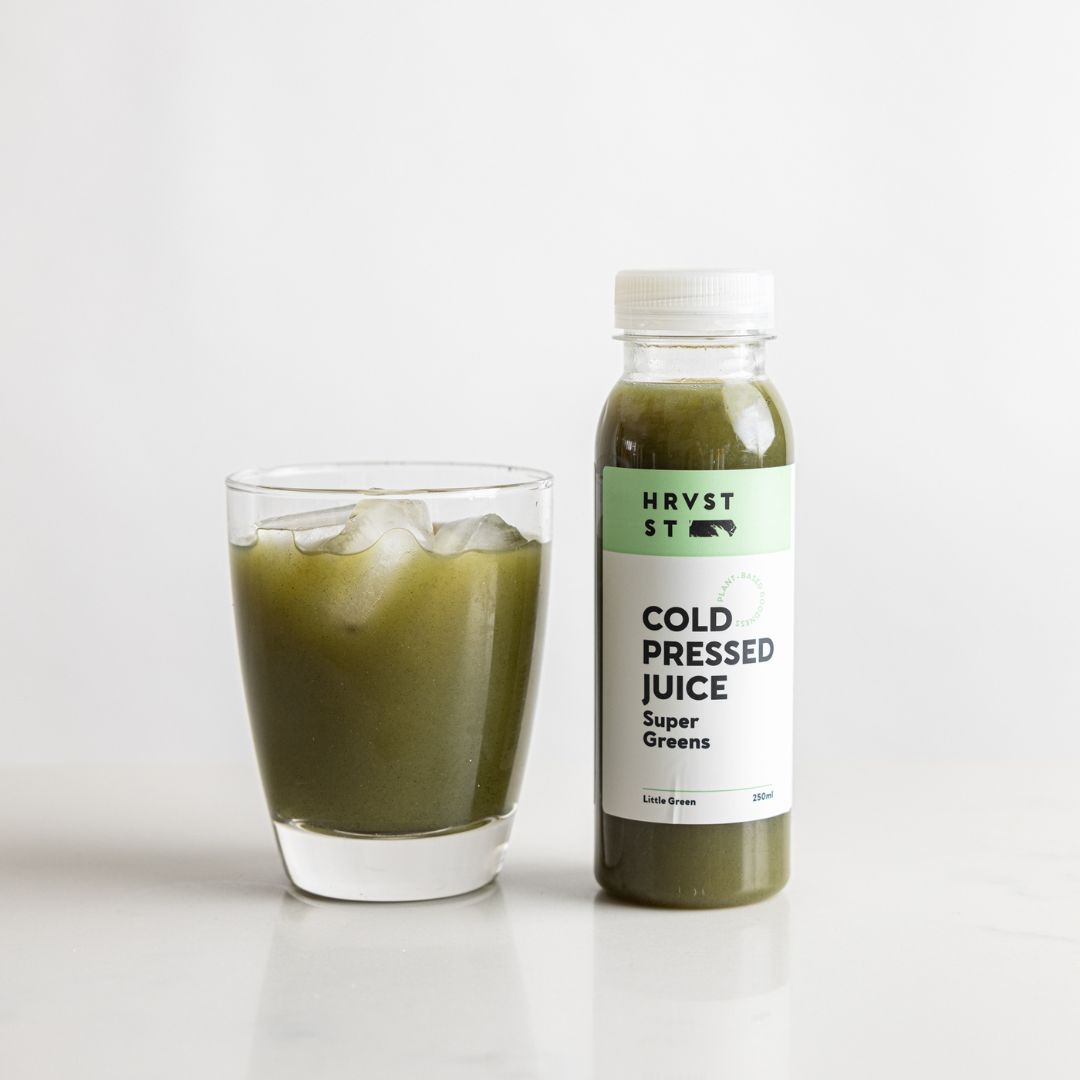 A drinking glass of green juice, served beside a bottled serve of green cold pressed juice, set against a white grey backdrop and bench.