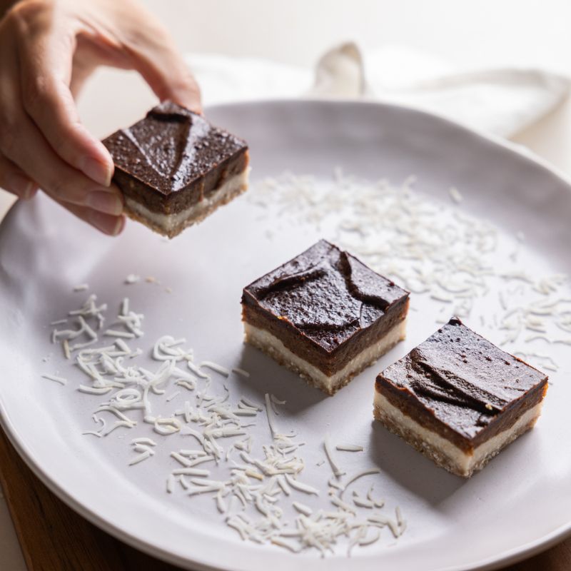 An action photograph of a hand picking up one of three salted caramel slices on a white plate. The plate has decorative sprinkles of dessicated coconut and is sitting on a white bench with a white cloth.