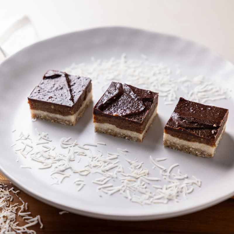 A photograph of three salted caramel slices serviced decoratively on a white plate with shreds of dessicated coconut on a white bench.