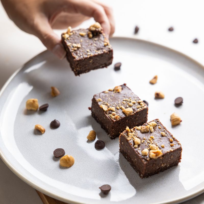An action photograph of a hand picking up one of three chocolate hazelnut slices off a white plate, decorated with chopped hazelnuts and chocolate chips.