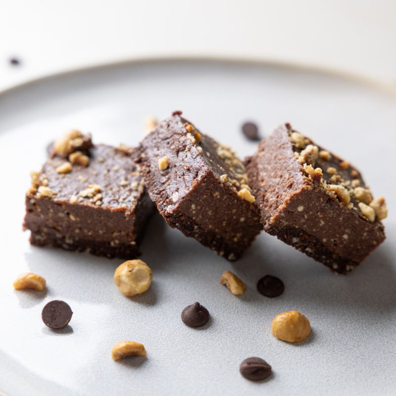 A photograph showing a side angle of three chocolate hazelnut slices leaning against each other and served on a white plate. A few chocolate chips and chopped hazelnuts decorate the plate, surrounding the slices.