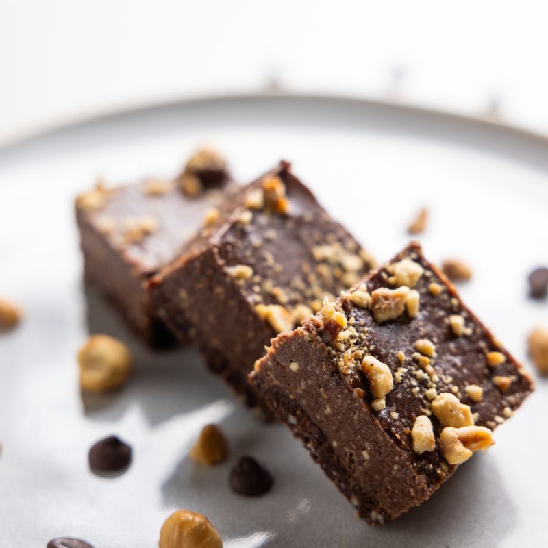 A close-up photograph of the top of a chocolate hazelnut slice leaning against two other slices. In the background is the white plate these slices are served on, chocolate chips, and chopped hazelnuts.