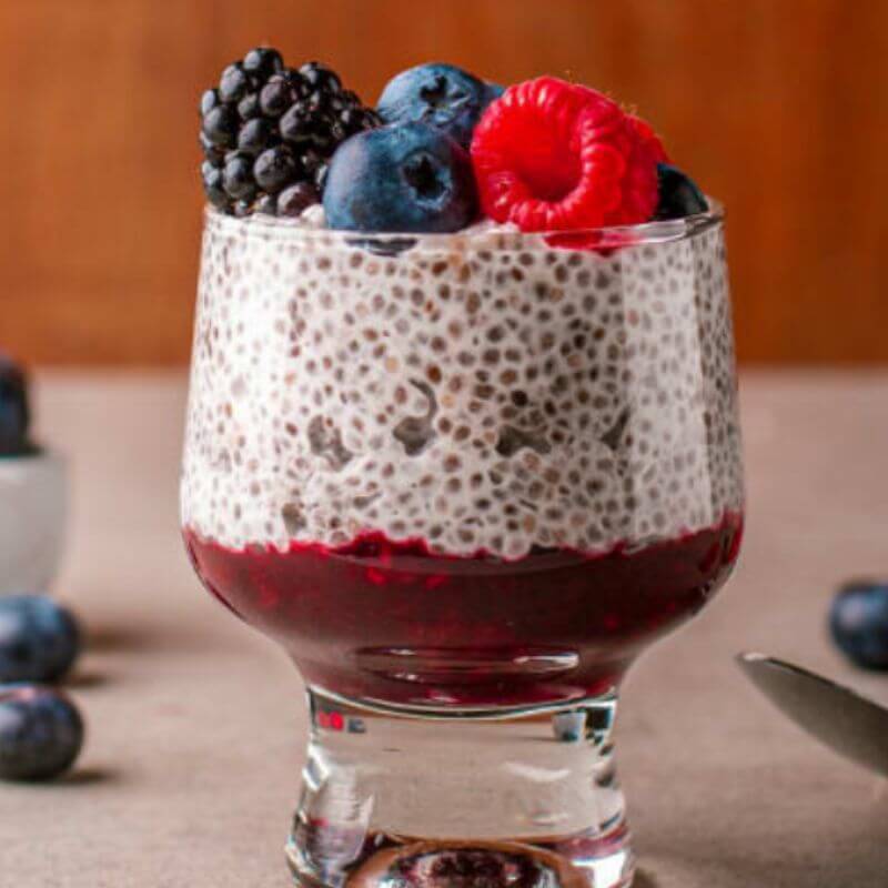 A small glass filled with layers of berry coulis, creamy chia pudding and fresh blackberries, blueberries and raspberries. The glass is sat on a marbel bench with a wooden backdrop, and some extra berries across the bench.