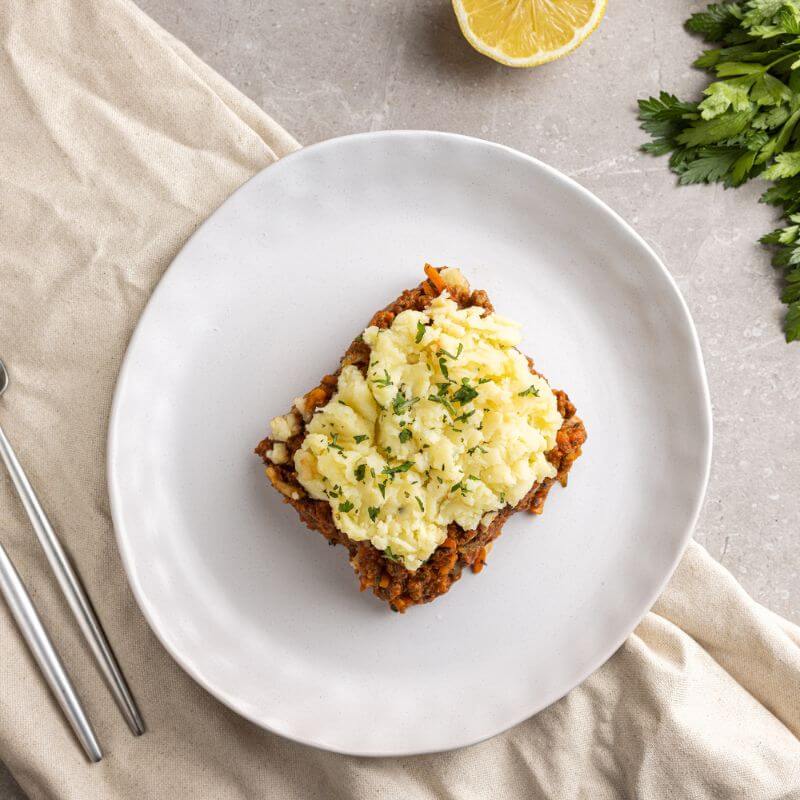 Flat lay image of Cottage Pie served on a white plate placed on a beige-colored cloth. A fork and knife sit neatly to the left of the plate. The Cottage Pie is garnished with freshly chopped parsley, adding a vibrant touch of green. In the background, a lemon and a bunch of parsley are subtly included for added freshness. The entire setup rests on a grey marble countertop, enhancing the warm and cozy aesthetic.