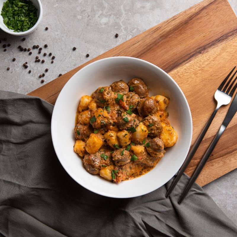 A top down photograph of an Italian Sausage Gnocchi served in a white bowl, set upon a wooden chopping board and dark grey cloth. A knife and fork sits to the left, and some peppercorns sit in a ramekin to the right.