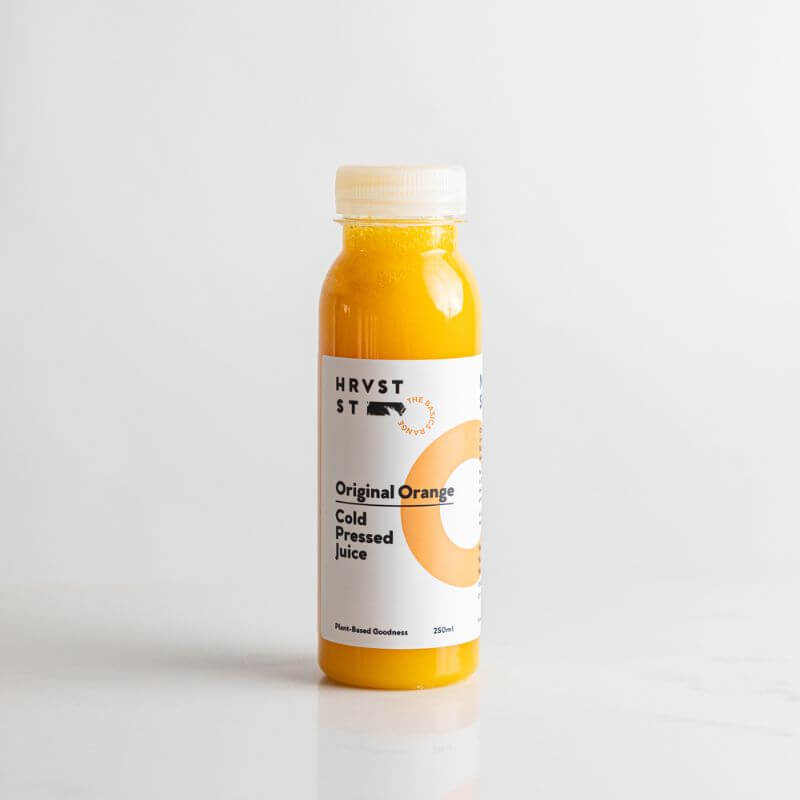 A full bottled serve of cold pressed orange juice set against a white grey backdrop and bench.