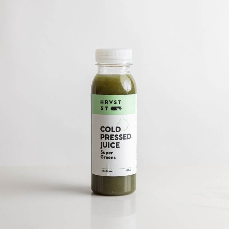 A full bottle of green cold pressed juice set against a white grey backdrop and bench.