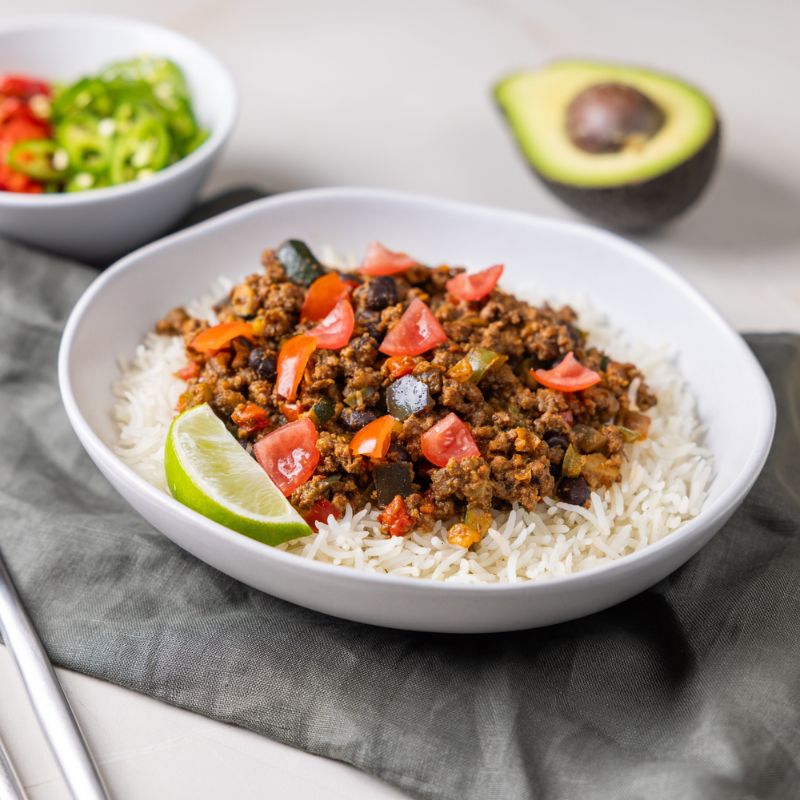 Side angle shot of the Burrito Bowl, showcasing its depth and layers. A white ramekin with chopped red and green capsicum and a sliced avocado are included in the frame for visual balance, reinforcing the Mexican-inspired theme.