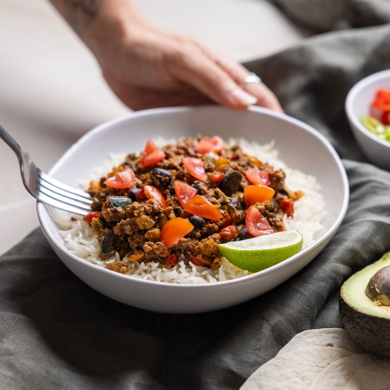 Action shot of one hand holding the Burrito Bowl while the other hand scoops up a piece of the seasoned beef with a fork. This adds a dynamic and inviting feel, making the dish look ready to be enjoyed.
