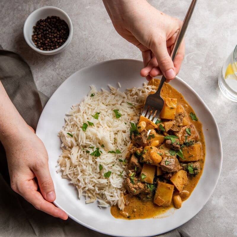 Action shot of the Beef Massaman, with one hand holding the plate and another hand using a fork to pick up a piece of beef. The shot conveys the dish's comfort and deliciousness.  "
