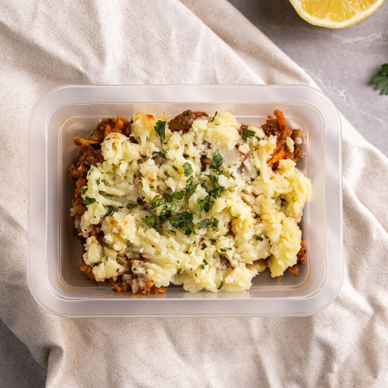 Clear container shot of the Cottage Pie, with its distinct mashed potato topping and beef filling visible. The container is placed on the beige cloth, with a fork and knife arranged neatly nearby. The background includes the grey marble countertop and a subtle hint of parsley to complete the presentation.