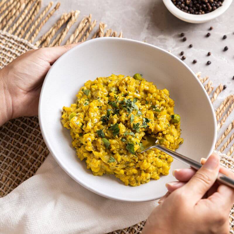 An action shot of Pumpkin Risotto, with one hand holding a white bowl and another hand using a fork to scoop up the creamy risotto. The dish has a smooth texture and is garnished with fresh chopped spring onion.