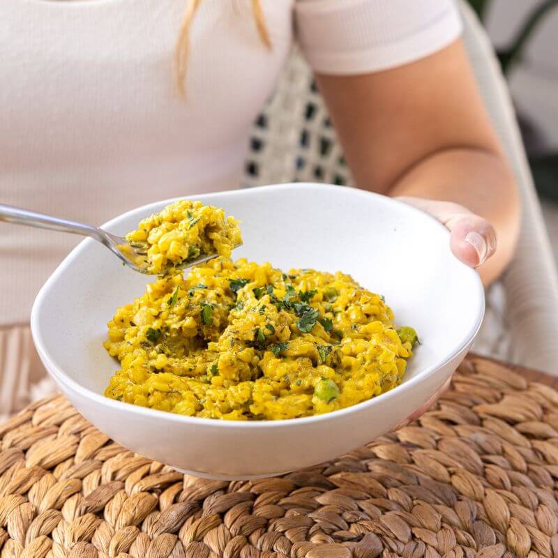 A clear plastic container of Pumpkin Risotto sits on a wool fringed placemat, with a white cloth tucked underneath. The vibrant golden risotto is smooth and creamy, with scattered black peppercorns adding a rustic touch to the shot.