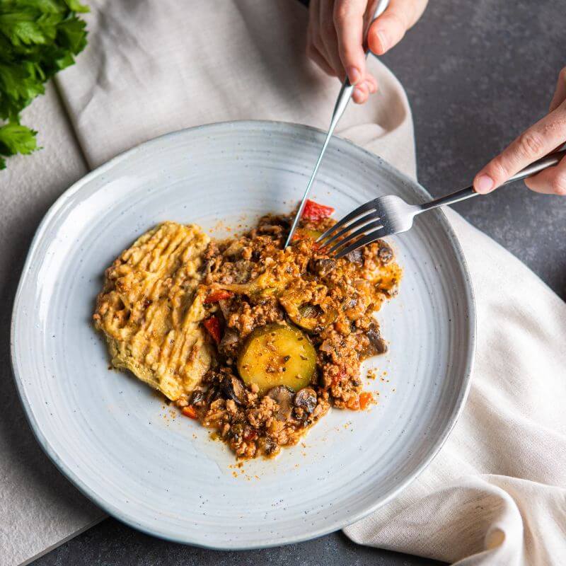 Action shot of two hands cutting into the Savoury Mince with Baba Ganoush using a fork and knife. The blue-hued plate and beige cloth remain visible, with parsley in the background adding a fresh, natural element.