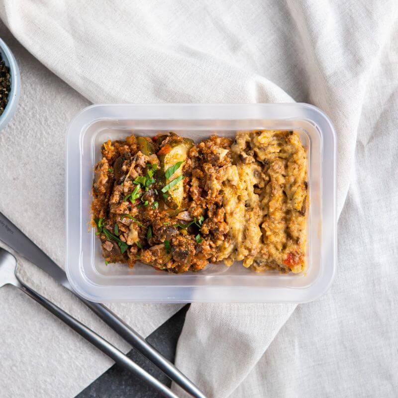 Clear container shot of the Savoury Mince with Baba Ganoush, emphasizing its portability and convenience. The fork and knife are positioned alongside the container, set on a beige cloth and dark marble background, with parsley scattered for garnish.