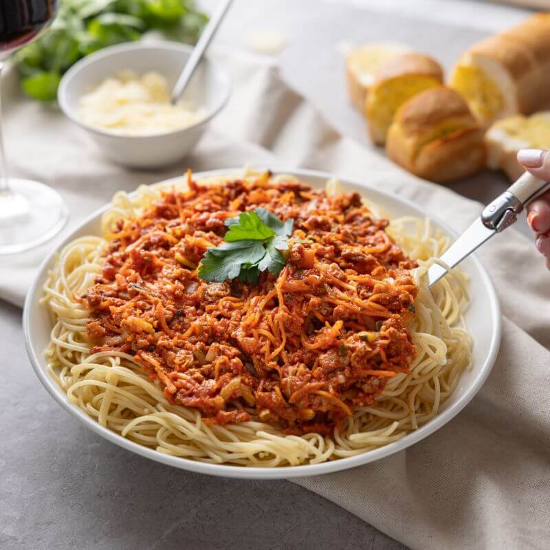 Action shot of a hand holding the serving spoon to scoop up Spaghetti Bolognese from the plate, with garlic bread, Parmesan cheese, and a glass of wine in the background creating a comforting dining scene.