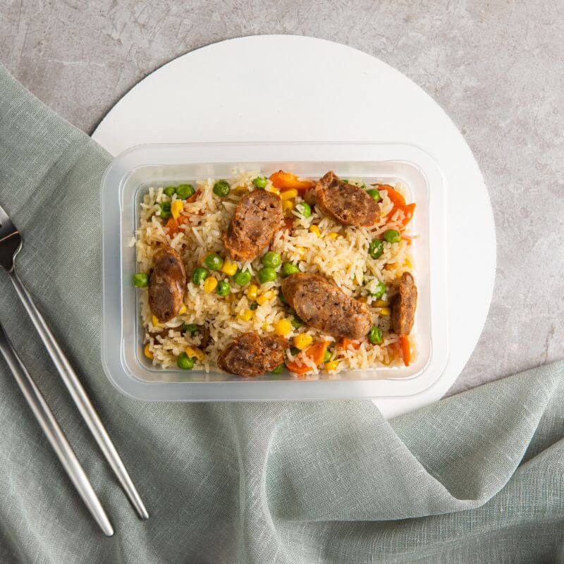 Our take on the classic Fried Rice served in a container, placed on a white circular placemat with a marble bench top and a teal coloured cloth underneath. A knife and fork sits to the left.