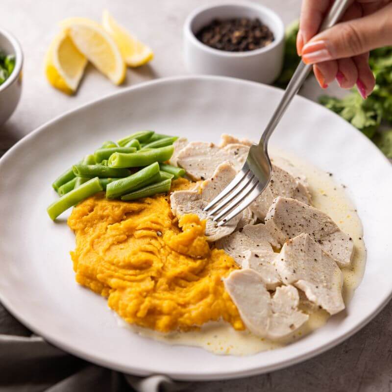 An action shot of Lemon Pepper Chicken, with a hand holding a fork that is scooping up a piece of chicken. The creamy lemon pepper sauce, green beans, and sweet potato mash are visible, alongside background elements such as sliced lemon wedges, a ramekin of cracked peppercorn, and a green cloth.
