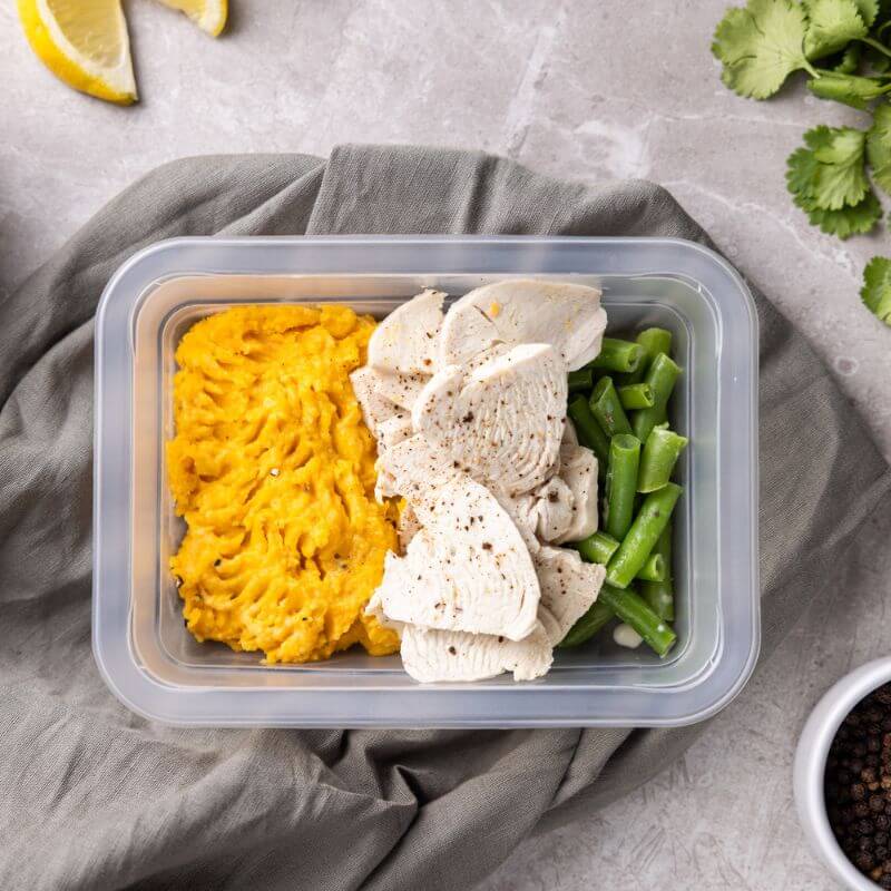 A clear plastic container of Lemon Pepper Chicken, featuring chicken breast in creamy lemon pepper sauce, green beans, and a bed of sweet potato mash. The container rests on a neutral background, showcasing the meal’s vibrant colors and textures.