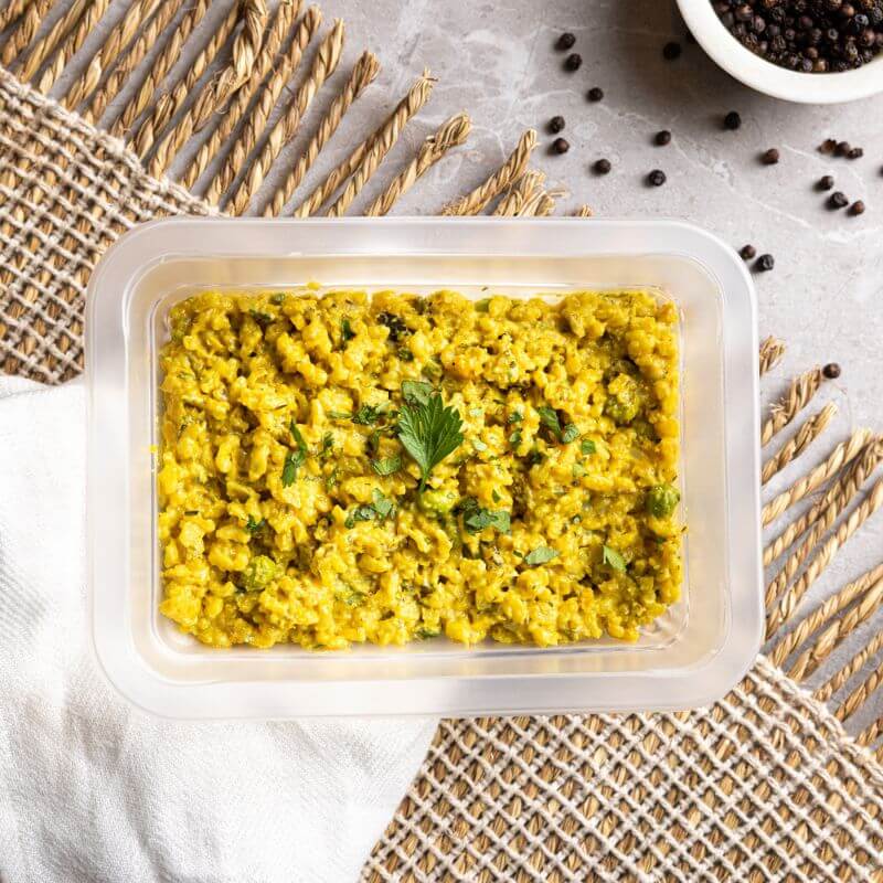 A clear plastic container of Pumpkin Risotto sits on a wool fringed placemat, with a white cloth tucked underneath. The vibrant golden risotto is smooth and creamy, with scattered black peppercorns adding a rustic touch to the shot.