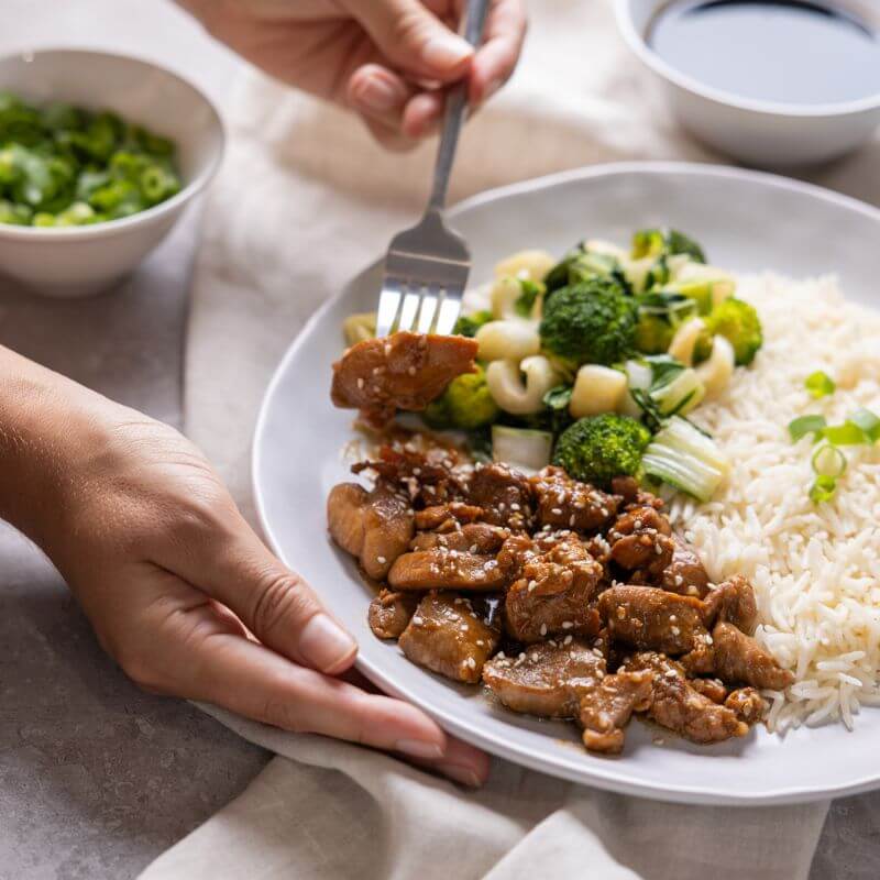 Action shot of two hands; one hand is holding the plate of Teriyaki Chicken, while the other is scooping up a portion of chicken. A white ramekin of chopped spring onion and gluten-free soy sauce are visible in the background.