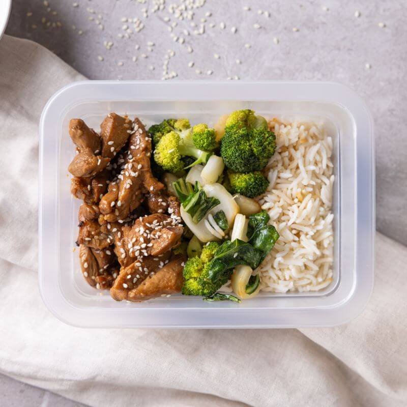 Clear container shot of Teriyaki Chicken, showing tender chicken, fluffy basmati rice, and steamed greens, garnished with sesame seeds for a convenient, ready-to-eat meal.