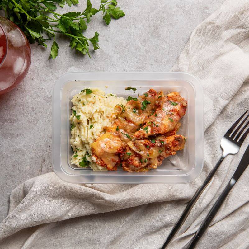A top down photograph of a cheese and tomato sauce covered chicken breast, served with a creamy white mashed potato. A knife and fork lay to the side, and the dish is served on a marble bench with a cream cloth underneath.