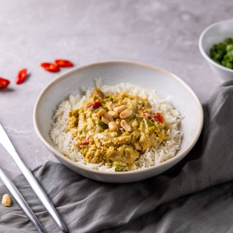 A side angle photograph of a ceramic bowl sitting on a grey marble bench and grey green cloth. The bowl contains basmati rice with a rich, satay chicken and peanuts sitting atop. Beside the bowl lays a knife and fork, some chopped chilli and ramekin of hopped spring onion.
