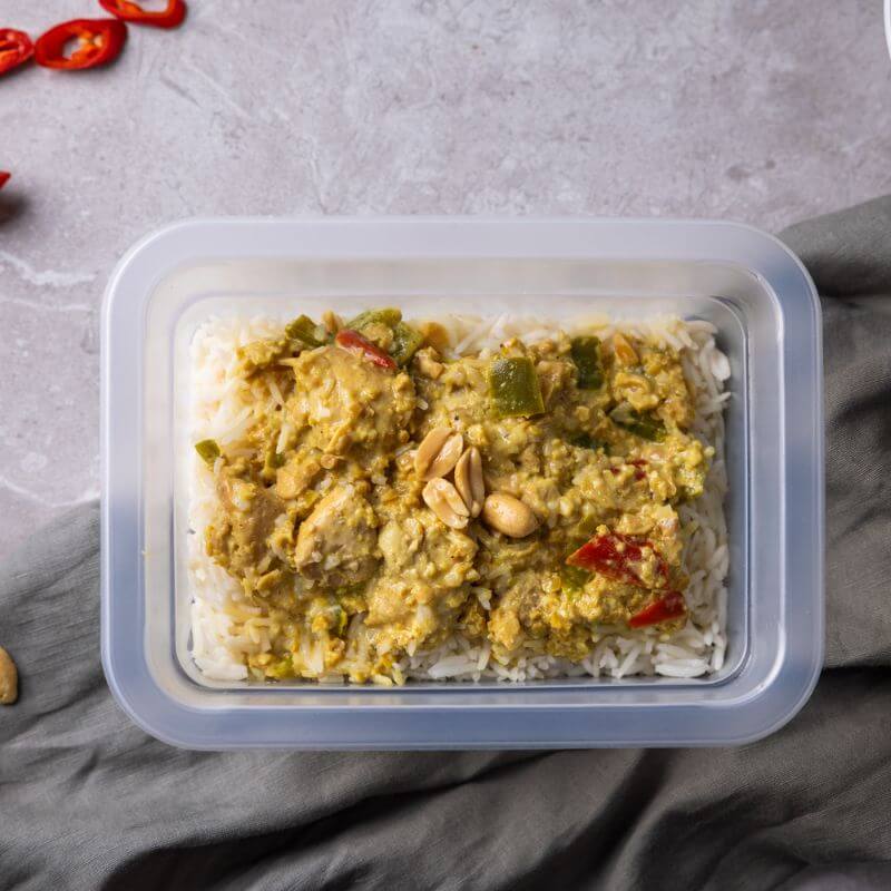 A top down photograph of a container filled with basmati rice and a rich, creamy satay sauce with chicken, capsicum and peanuts. The container is placed on a grey marble bench top with a green grey cloth beneath it.