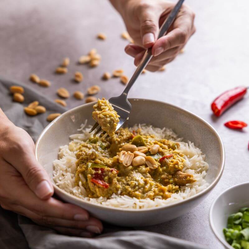 A photograph of a hand holding a white ceramic bowl set on a marble bench top and a green grey cloth beneath. The bowl is filled with basmati rice and rich satay chicken, and a hand is holding a fork over the bowl, scooping out a piece of chicken. Surrounding the bowl are some peanuts, chilli and spring onions.