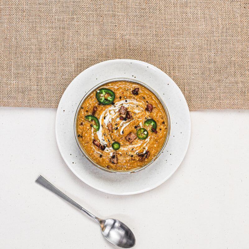 Flat lay image of the Bacon & Jalapeño Soup served in a white bowl. The bowl is placed on a white plate, atop a beige rattan placemat on a white countertop. A silver spoon is visible in the frame, completing the warm and inviting setup.