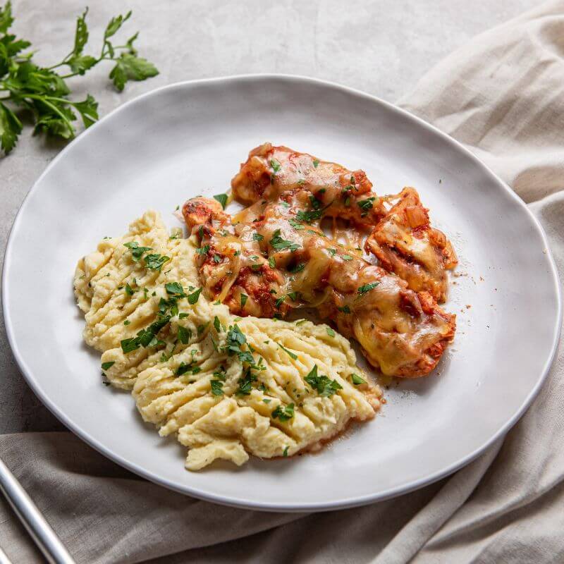 A side on photograph of chicken, covered in a rich tomato sauce and melted cheese, served on a white plate with creamy white mashed potato. The plate sits upon a white cloth and marble bench top, and there are some fresh green herbs in the background.